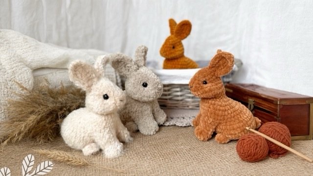 Three handmade crocheted baby rabbits displayed with yarn, a crochet hook, and a basket